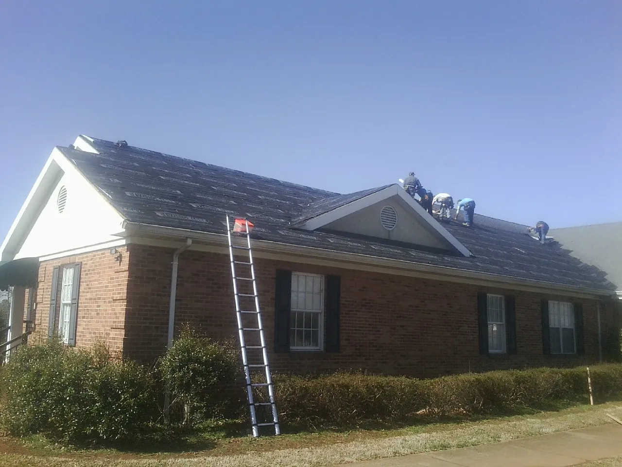Crew tearing shingles off a brick ranch roof with a ladder set — Metal Roof Repair work by our crew in Roberta