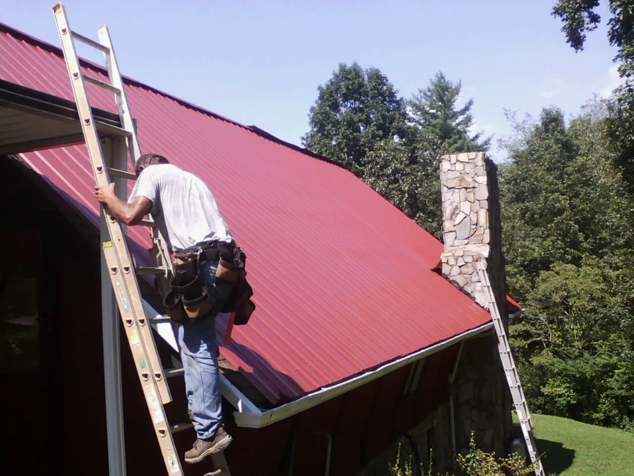 Roofer on a ladder beside a red metal roof and stone chimney — Soffit & Fascia Repair by Roofing Brothers in Roberta